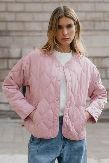 Woman wearing a pink quilted jacket and blue jeans against a brick wall.