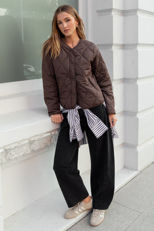 Woman standing against a white building wearing a brown quilted jacket and black pants.