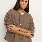 Woman wearing an oversized quilted mocha colored jacket with button front, styled with blue jeans and gold hoop earrings.