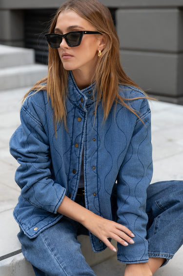 Woman wearing a blue denim jacket and sunglasses sitting on steps.