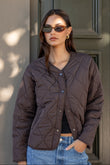 Woman wearing a brown quilted jacket and sunglasses standing against a neutral wall.