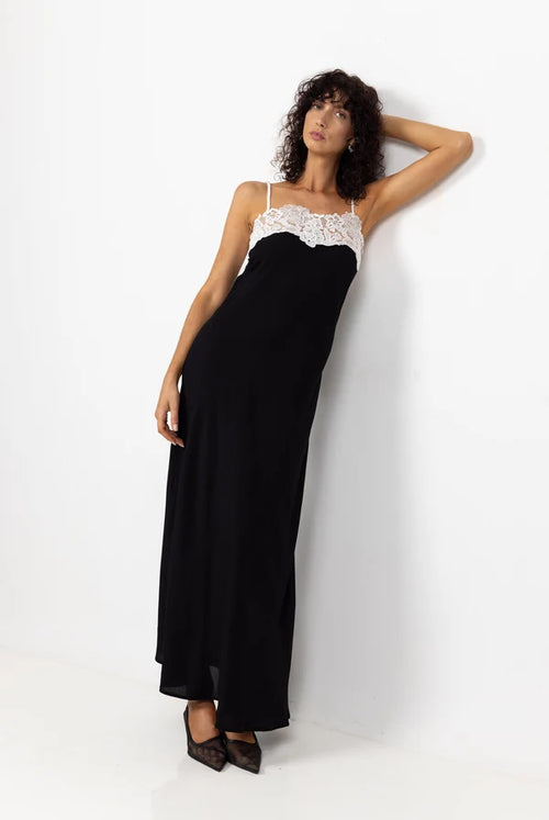 A woman modeling a black maxi dress with a white lace-trimmed spaghetti strap bodice, leaning against a white wall.