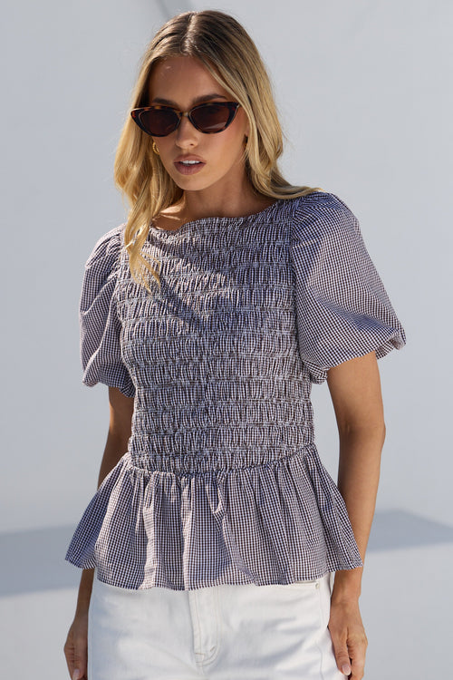 A woman modeling a smocked short-sleeve blouse with a blue and white gingham pattern, paired with white pants, set against a clean white background.
