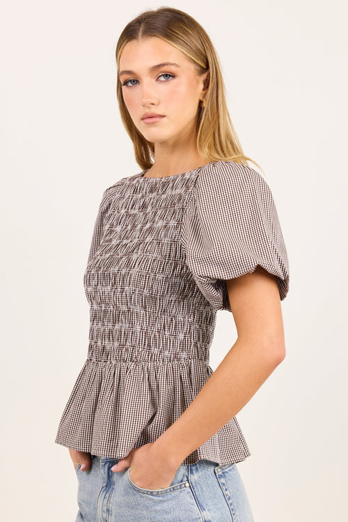 A woman modeling a short-sleeve smocked blouse with a brown and white gingham pattern, paired with light-wash blue jeans, against a plain off-white background.