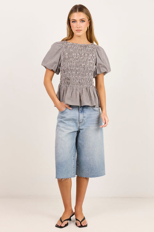 A woman modeling a light gray smocked puff-sleeve top paired with light-wash denim culottes and black sandals, standing against a plain off-white studio background.