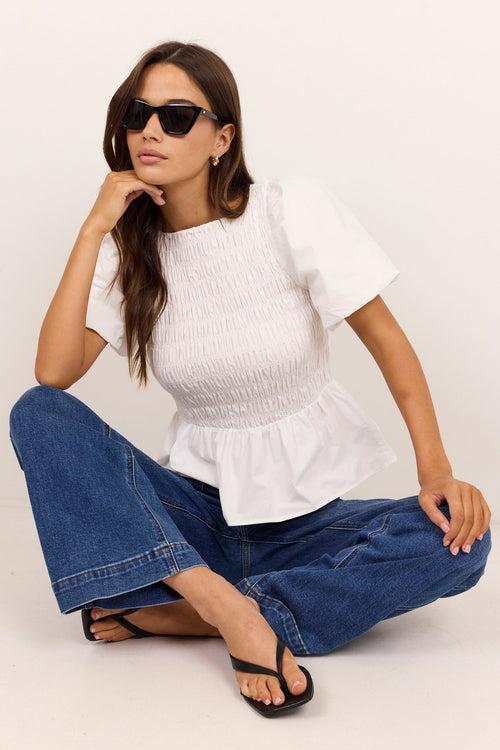 A woman sits cross-legged on a white studio floor wearing a white smocked short-sleeve peplum top, blue wide-leg jeans, and black flip-flops.