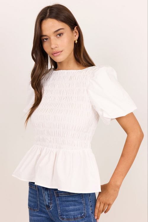 A woman wearing a white smocked blouse with puff sleeves and a peplum hem, paired with blue jeans, against a plain off-white background.