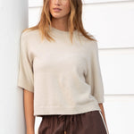 Woman wearing a beige top and brown pants leaning against a white wall.