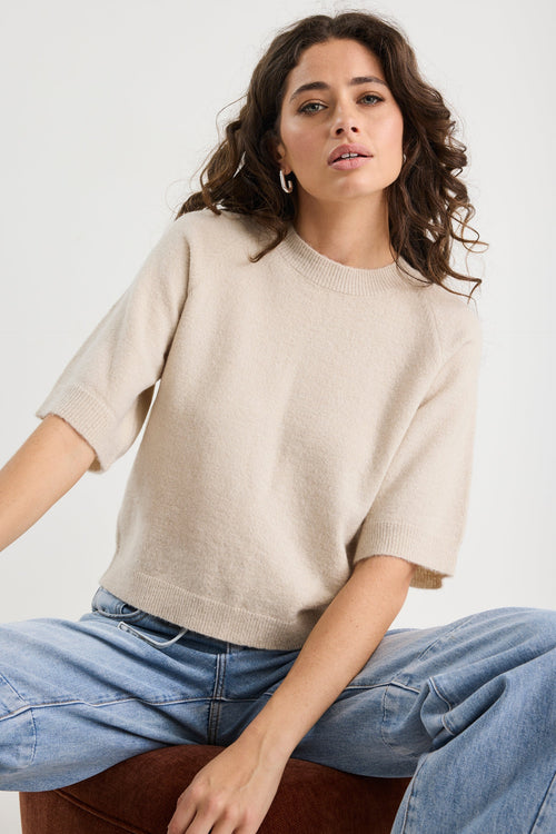 Woman wearing a beige sweater and blue jeans sitting on a white background
