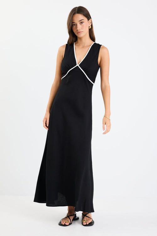 Woman wearing a black sleeveless maxi dress with a V-neckline, white contrast binding detail, bias cut, and softly flared hem.