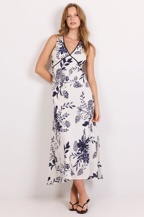 A woman modeling a sleeveless midi dress with a navy blue floral print on a white background, standing against a plain white studio backdrop.