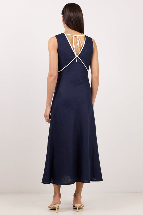 A woman stands with her back to the camera wearing a sleeveless navy midi dress with a white crisscross lace-up back detail, paired with beige heeled sandals, against a plain white studio background.
