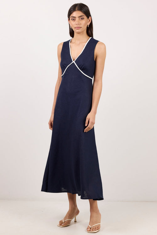 A woman modeling a sleeveless navy maxi dress with white contrast trim along the V-neckline and waist, paired with clear high-heeled sandals, against a plain off-white studio background.