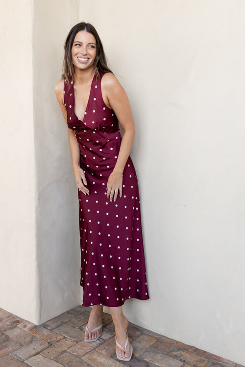 A woman wearing a sleeveless burgundy midi dress with white polka dots, standing against a beige stucco wall on a brick patio.