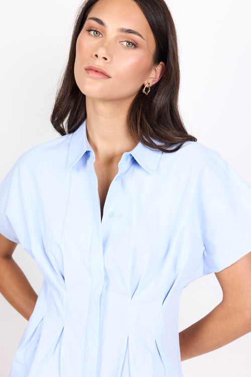 A woman wearing a light blue short-sleeve button-up shirt dress, photographed against a clean white studio background.