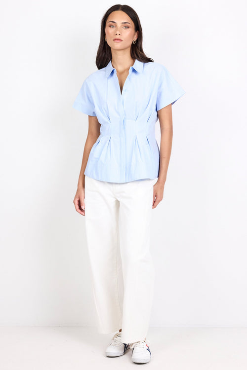 A woman modeling a light blue short-sleeve button-up blouse with a gathered waist and white wide-leg trousers, standing against a plain white studio background.