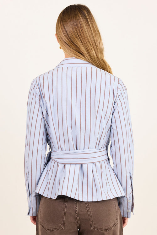 A woman viewed from behind wearing a light blue and white vertically striped belted jacket and brown pants, set against a plain off-white background.