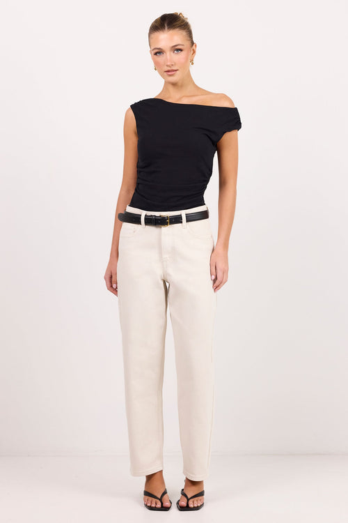 A woman modeling a black off-the-shoulder short-sleeve top and cream-jeans with a black belt, standing against a plain white studio background.