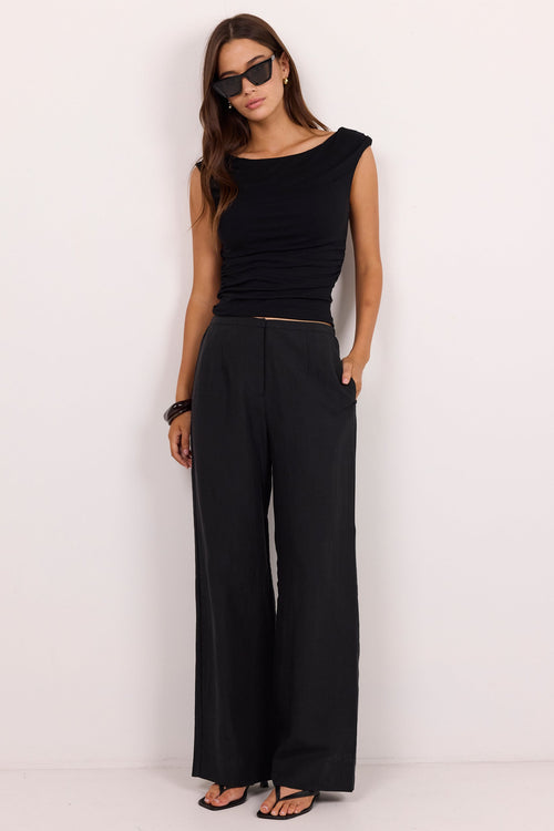 A woman modeling a black sleeveless top and black wide-leg trousers, standing against a plain white studio wall.