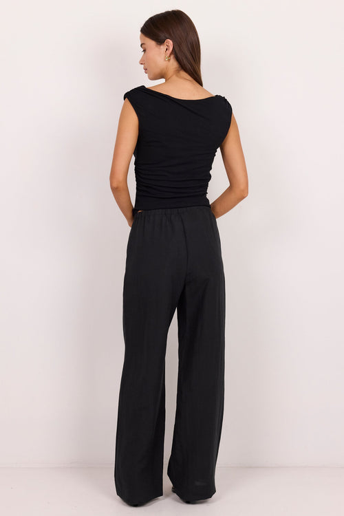 A woman is shown from the back wearing a black sleeveless top and matching black wide-leg trousers, standing against a plain white studio background.
