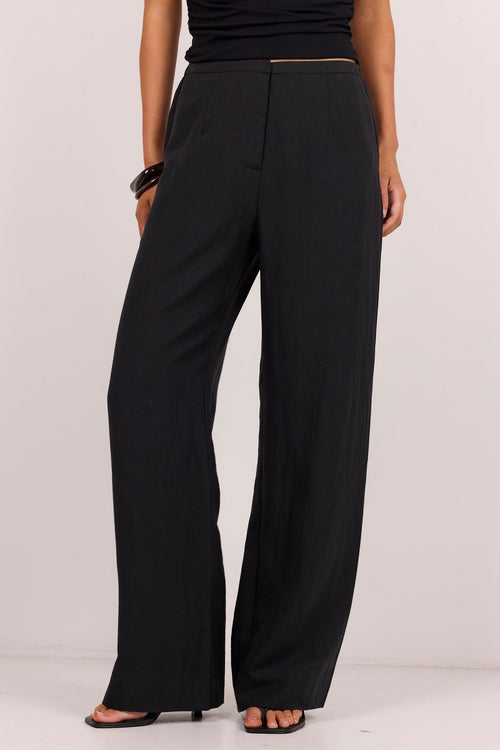 A model wearing black wide-leg trousers with a black sleeveless top and black sandals, shown from the waist down against a plain light gray studio background.
