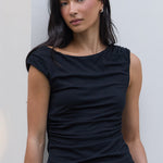 A woman wearing a black sleeveless top and brown shorts with a black belt, standing against a white building wall.
