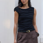 A woman stands outdoors against a white wall wearing a black sleeveless top and brown pleated shorts with a black belt. She is holding a black handbag.
