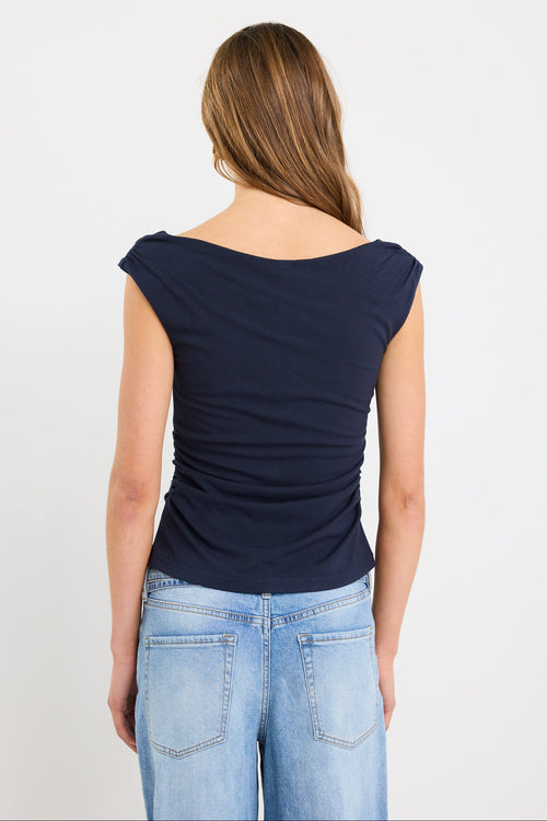 Woman wearing a fitted navy sleeveless top with boat neckline and side ruching, paired with light blue jeans.