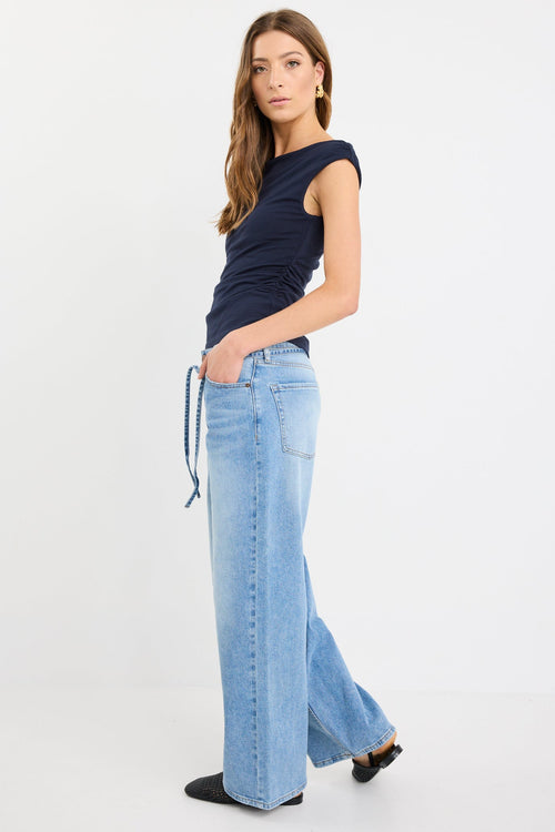 Woman wearing a fitted navy sleeveless top with boat neckline and side ruching, paired with light blue jeans.