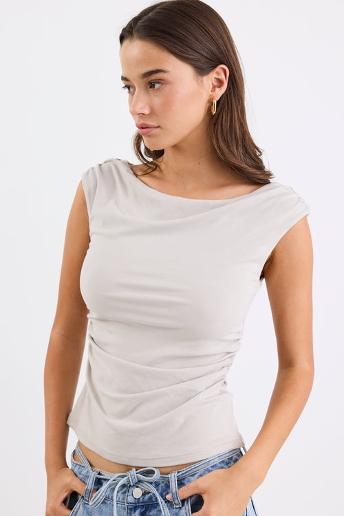 Woman wearing a stone-coloured sleeveless stretch top with a boat neckline, side ruching, and a fitted silhouette, paired with light blue jeans.