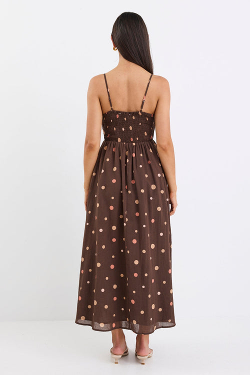 Woman wearing a chocolate brown midi dress with pink and beige polka dots, featuring a pleated bodice, thin straps, and gathered waist, styled with nude heels against a plain white background.