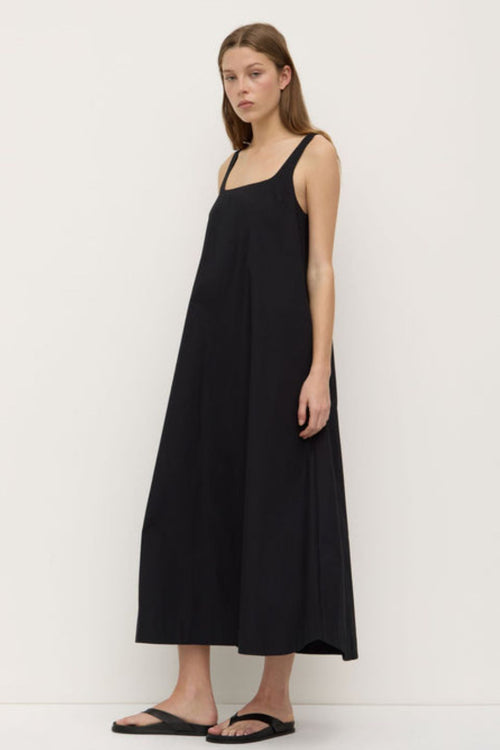 A woman wearing a black sleeveless maxi dress with wide straps and a square neckline. The dress has a flowing A-line silhouette and is styled with simple black sandals.