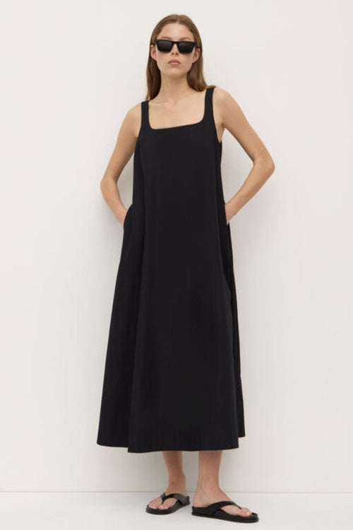 A woman wearing a black sleeveless maxi dress with wide straps and a square neckline. The dress has a flowing A-line silhouette and is styled with simple black sandals.