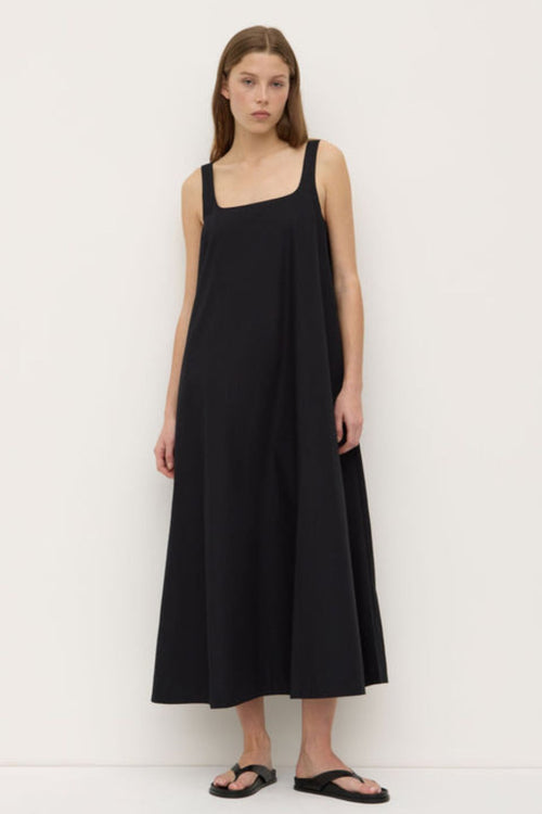 A woman wearing a black sleeveless maxi dress with wide straps and a square neckline. The dress has a flowing A-line silhouette and is styled with simple black sandals.