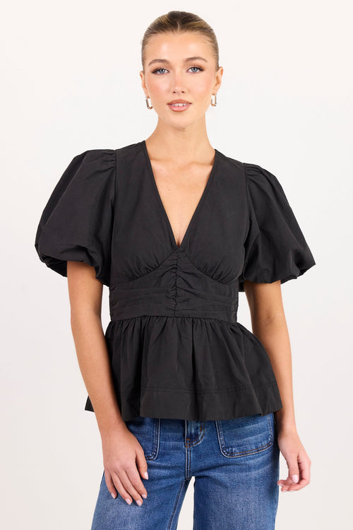 A woman modeling a black puff-sleeve top with a deep V-neck and gathered peplum waist, paired with blue jeans, against a plain white studio background.