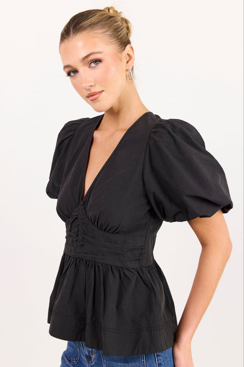 A woman wearing a black puff-sleeve peplum top with a deep V-neck and gathered waist, paired with blue jeans, photographed against a plain light background.