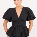 A woman modeling a black puff-sleeve peplum top with a deep V-neck and gathered waist, paired with blue jeans, against a plain light background.