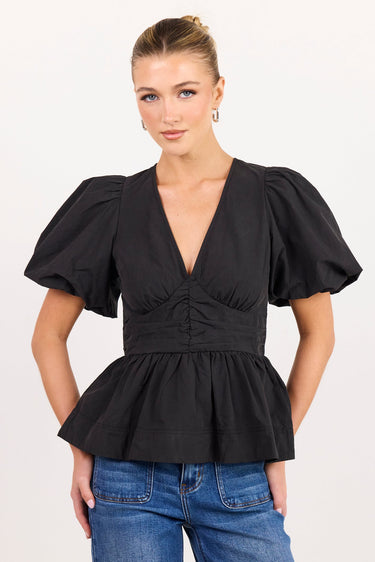 A woman modeling a black puff-sleeve peplum top with a deep V-neck and gathered waist, paired with blue jeans, against a plain light background.