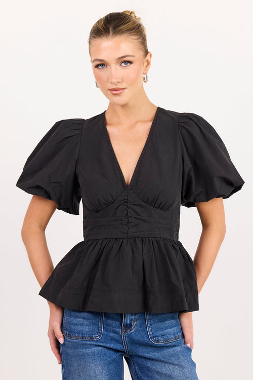 A woman modeling a black puff-sleeve peplum top with a deep V-neck and gathered waist, paired with blue jeans, against a plain light background.