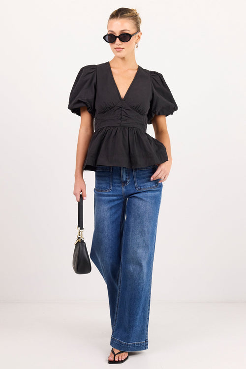 A woman modeling a black short-sleeve peplum blouse with puff sleeves, paired with wide-leg blue jeans, black sunglasses, and a small black handbag, against a plain light gray studio background.