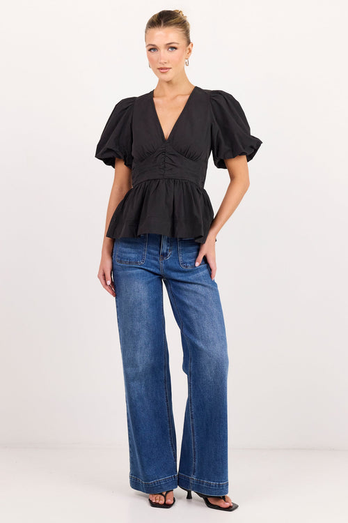 A woman modeling a black short-sleeve peplum top with puff sleeves and blue wide-leg jeans against a plain white studio background.