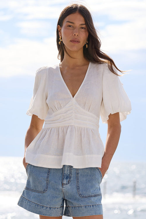 A woman stands on a concrete pier by the ocean wearing a white short-sleeve peplum-style top, light-wash denim shorts, and dark sandals under a bright sky.