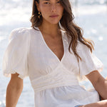 A woman sits on a concrete pier by the ocean, wearing a white puff-sleeve wrap top, denim shorts, and black sandals.