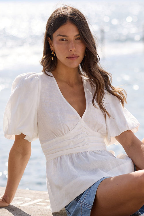 A woman sits on a concrete pier by the ocean, wearing a white puff-sleeve wrap top, denim shorts, and black sandals.