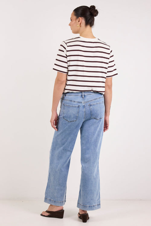 A woman is shown from the back wearing a black and white horizontally striped short-sleeve t-shirt, light blue wide-leg jeans, and dark brown wedge sandals, standing against a plain white studio background.