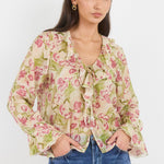 A woman modeling a long-sleeve floral blouse with a tie-neck detail and ruffled cuffs, paired with blue jeans, against a plain light gray background.