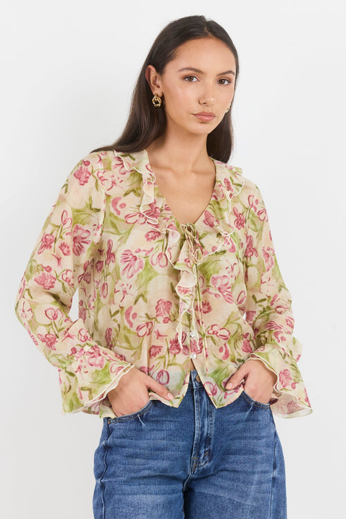 A woman modeling a long-sleeve floral blouse with a tie-neck detail and ruffled cuffs, paired with blue jeans, against a plain light gray background.