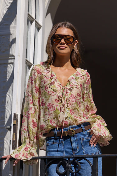 A woman wearing a beige floral long-sleeve blouse with tie-front detailing, blue jeans, and sunglasses, standing on a balcony with white architectural columns.