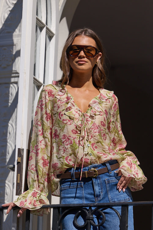 A woman wearing a beige floral long-sleeve blouse with tie-front detailing, blue jeans, and sunglasses, standing on a balcony with white architectural columns.