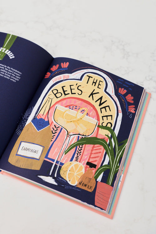 An open illustrated book or magazine spread on a light marble surface, featuring the title 'THE BEE'S KNEES' with a stylized cocktail glass, champagne bottle, lemon, and potted plant on a dark navy background.